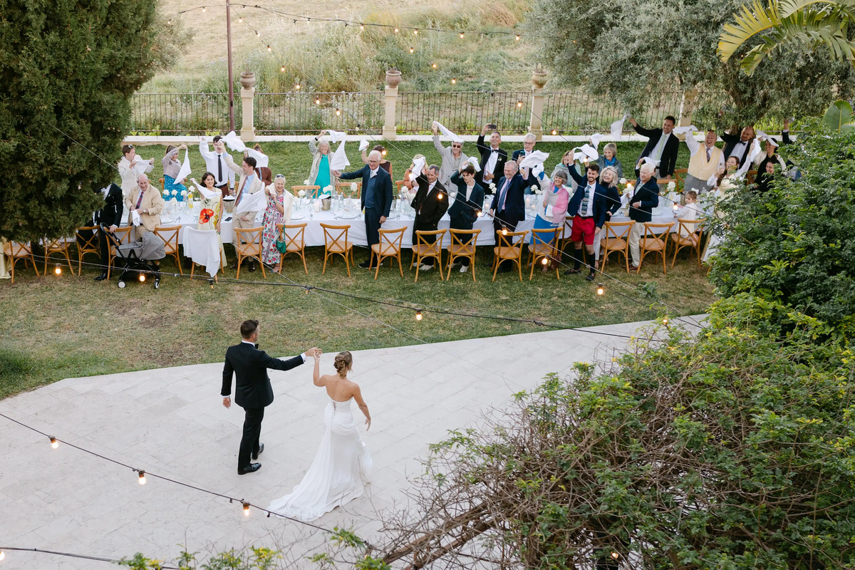 Bride and groom welcomed by guests waving napkins during an outdoor wedding dinner in Italy.
