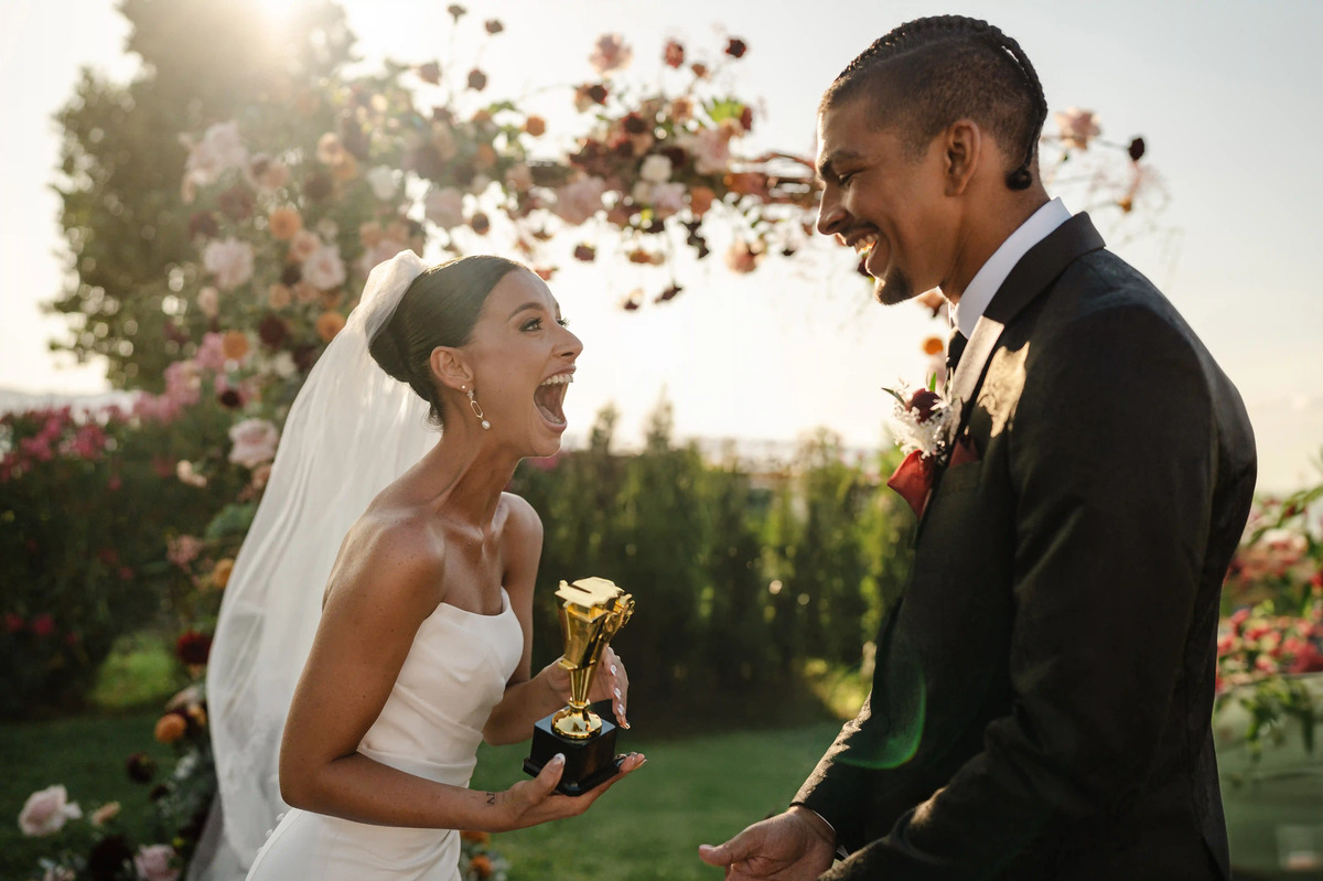 Bride in a strapless wedding dress laughing joyfully while holding a small golden trophy, facing the groom during an outdoor ceremony at sunset with floral arch in the background.